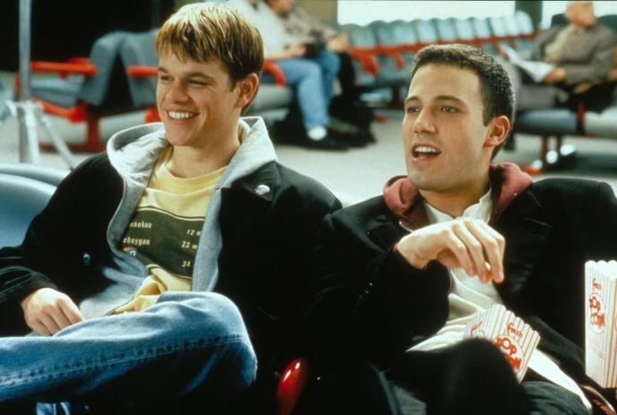 Airport lounge with two people seated smiling, one eating popcorn, red chairs, other passengers visible in the background.