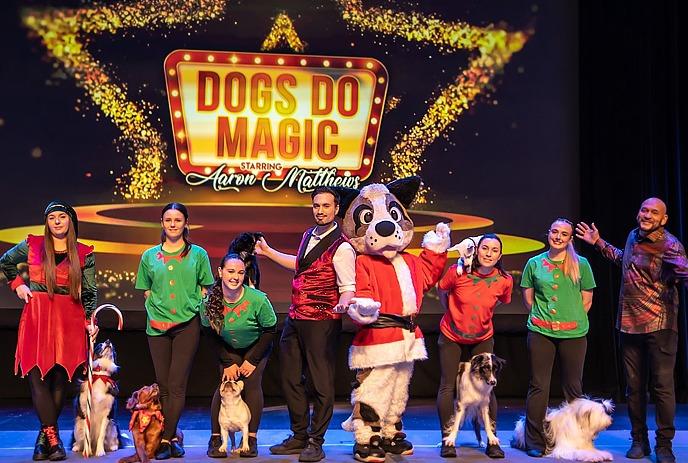 Dog mascot in silver jacket stands between two performers in matching silver outfits and magic sticks.