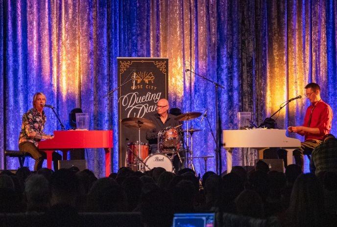 Musicians perform onstage with two pianos and drums before an audience in a lively concert setting.
