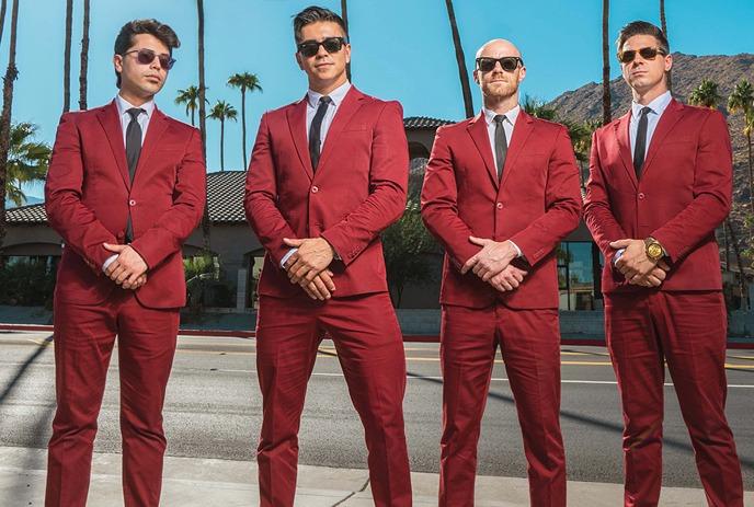 Four band members dressed in red suits, standing on a boulevard with palm trees in the background.