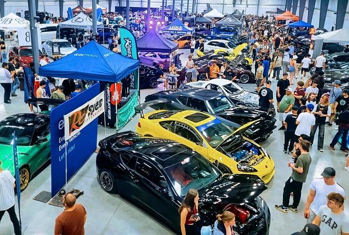 Crowds walk through a bright indoor car show filled with colorful modified cars displayed under tall lights