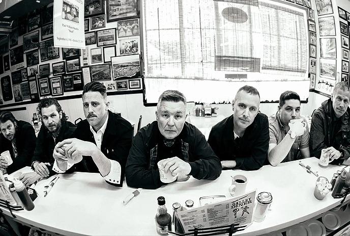Seven band members seated at a diner counter in a black-and-white wide-angle photo.