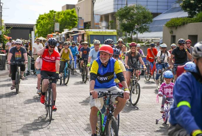Cyclists ride together in bright community event with helmets colorful outfits trees buildings festival motion.