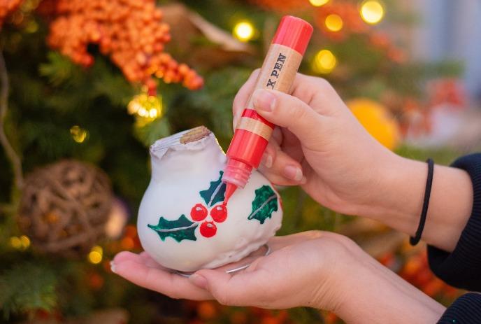 Hand decorates ceramic ornament with red pen; holly leaves and berries, Christmas tree behind.