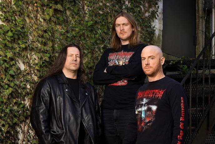 3 band members of Dying Fetus posing in front of a leaf covered wall.