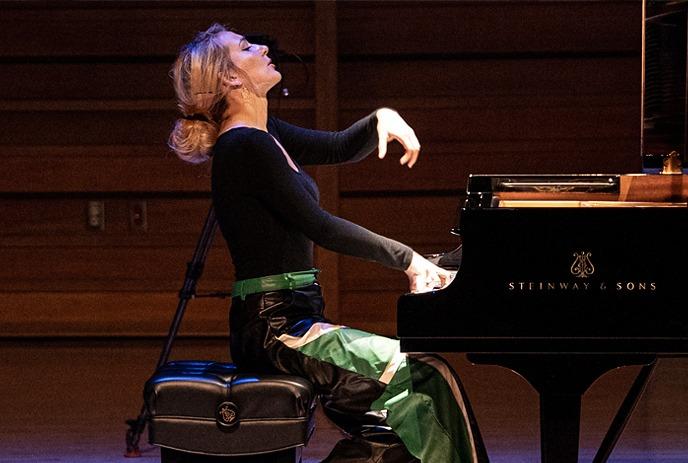 Pianist performing on grand piano in wood stage hall wearing black top and green pants focused on playing.