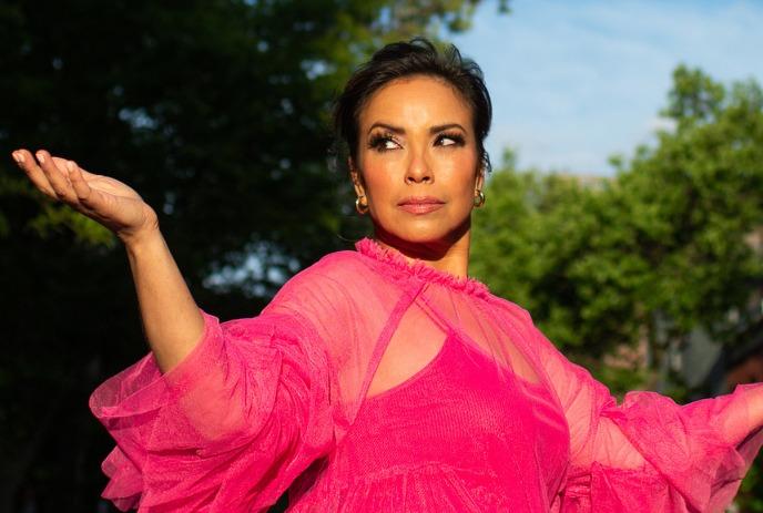 Emm Gryner in a vivid pink dress raises arms joyfully on a cobblestone street, framed by parked cars and leafy trees.