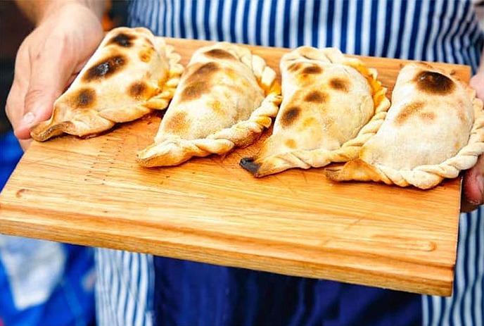 Three vendors in aprons serve empanadas at an outdoor stall beside a bright Colombian flag.