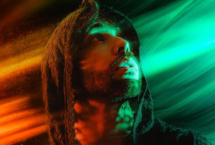 Ero808 in a hood gazes upwards, enveloped in swirling red and green light effects.