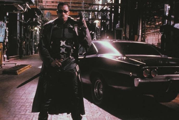 A warrior in black leather wields a sword, standing confidently beside a sleek muscle car in an industrial setting.