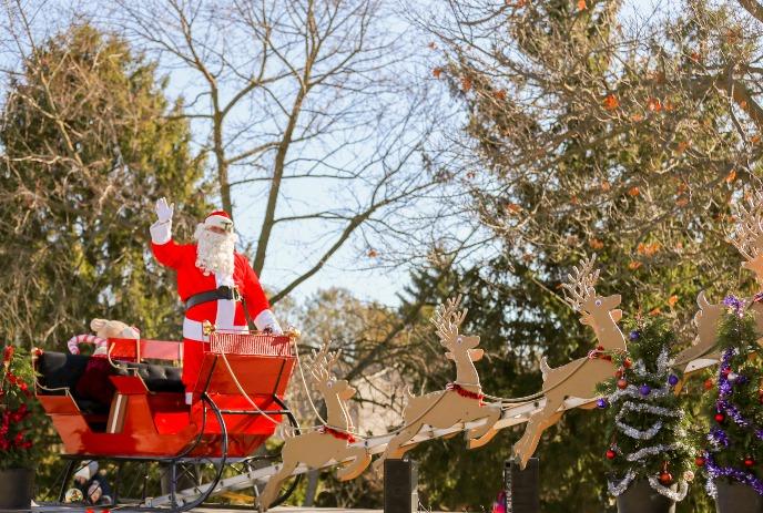 Santa stands on a red sleigh being pulled by reindeer and waves to the crowd.