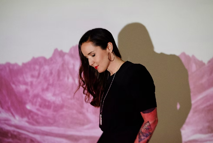 Sarah MacDougall looking down at the ground, with light casting her shadow on the wall behind her.