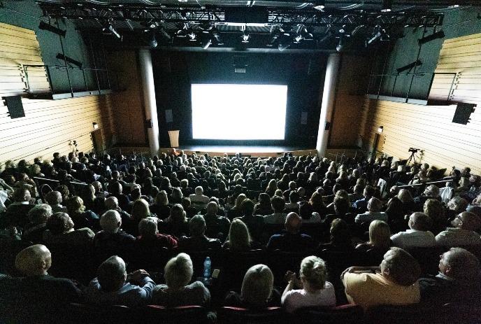 A large audience is seated in a theater, facing a brightly lit screen.