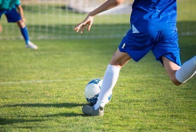 Player in blue kicks a soccer ball on grass as another player waits near the goal in the background.