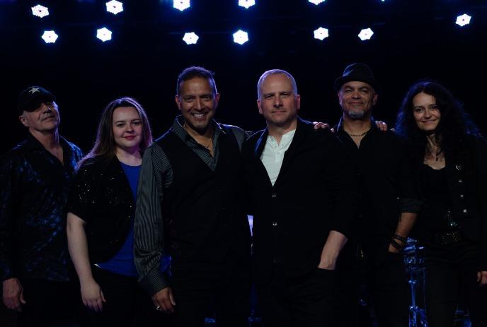 Six artists in dark outfits pose under rows of bright lights with a smile.