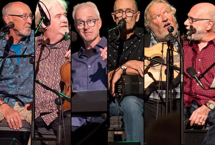 Six performers seated on stage each behind microphone with varied instruments illuminated by focused lights.