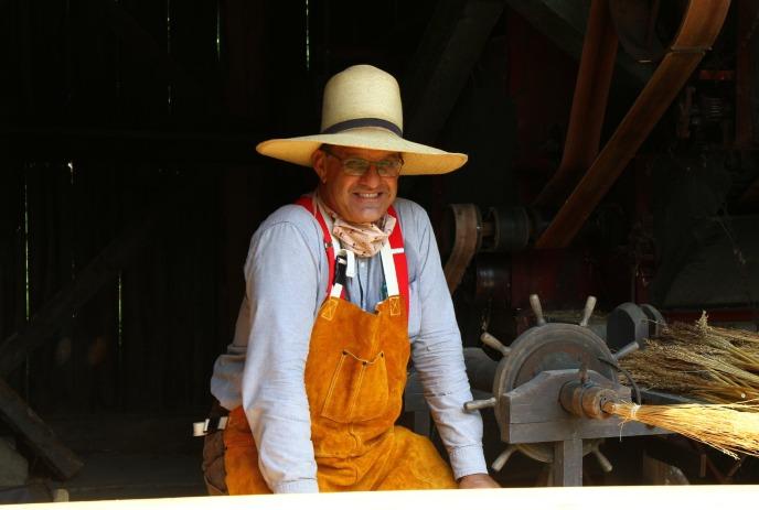 A person in a straw hat and leather apron stands in a barn by a threshing machine with straw.