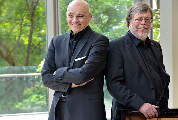 Leslie Kinton and James Anagnoson wearing black suites, posing for the camera in front of trees on a balcony.