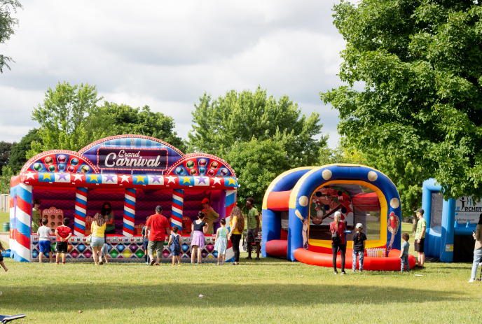 A colorful carnival scene with inflatable game booths, joyful people, lush green trees, and a cloudy sky.