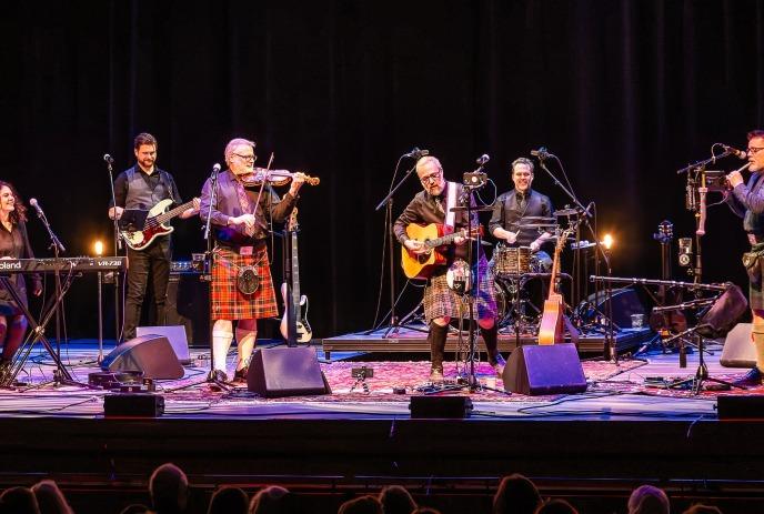 Band performing live with kilts, bagpipes, guitars, drums in a spotlight.