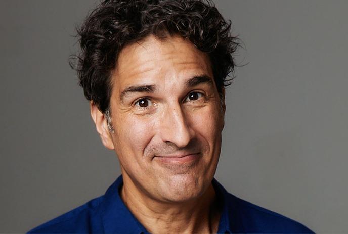 Gary Gulman smiles with a playful expression, wearing a blue button-up shirt, in gray studio background.