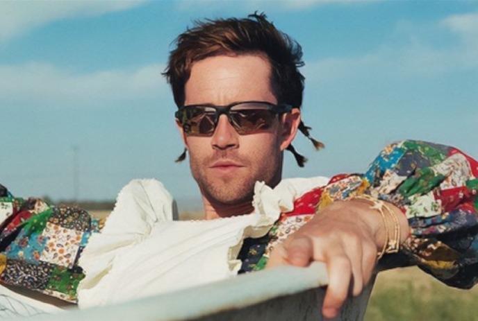 Geordie Kieffer wears sunglasses and a colorful patchwork outfit while leaning forward outdoors under a blue sky.