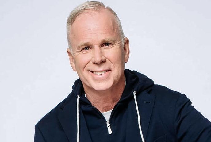 Gerry Dee sits wearing a blue hoodie under a blue sport coat and smiling at the camera.
