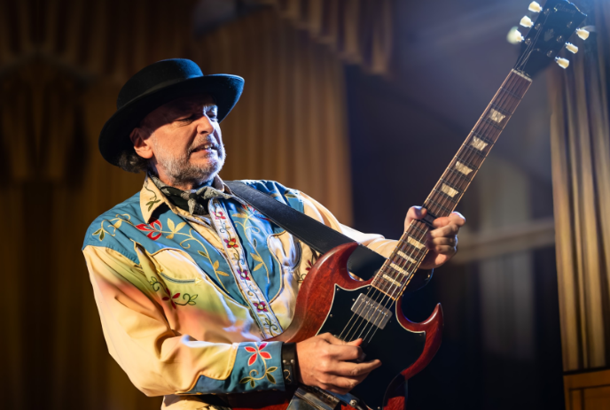 Musician in colorful embroidered shirt playing red electric guitar before golden curtains in warm lighting.