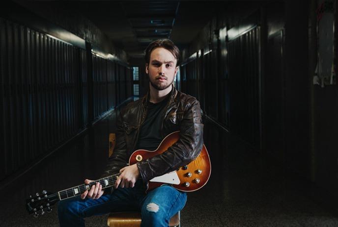 Spencer Mackenzie with a serious expression, sits in a dimly lit hallway, holding a sunburst electric guitar.