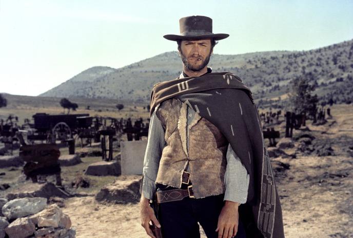 A cowboy in a desert landscape, dressed in a hat, poncho, vest, and belt with mountains behind.