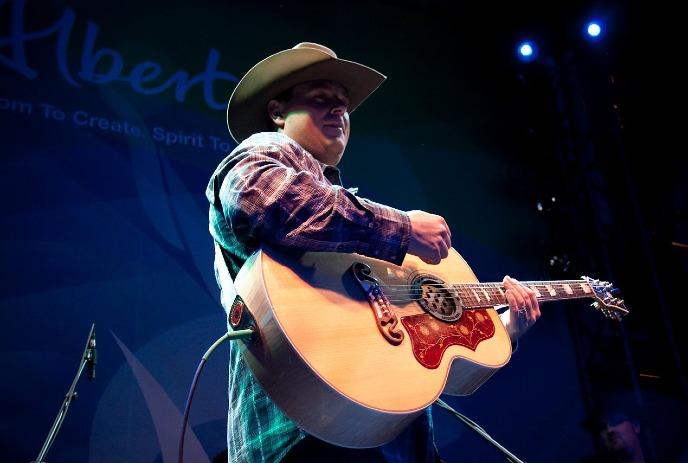 Cowboy?hatted musician in plaid plays acoustic guitar on lit stage with blue purple concert glow.