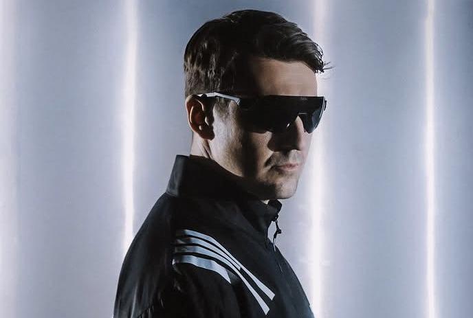 Artist in sunglasses against bold hexagon backdrop creating a sleek modern portrait aesthetic.