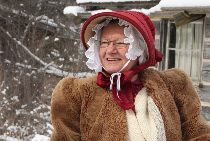 Lady in red bonnet and fur coat stands outside snowy cabin on a cold winter day.