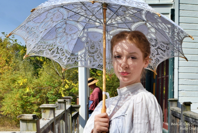A woman in vintage white attire holds a lace parasol outdoors.