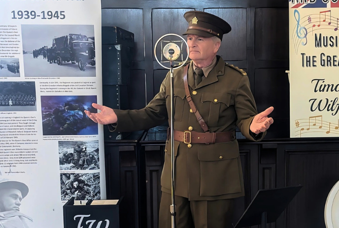 Uniformed speaker standing before wartime display panels giving a presentation in a dim wood interior setting.