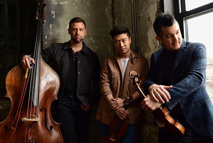 Three artists stand together with their instruments while looking at the camera.