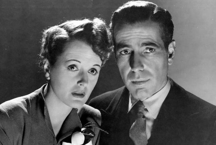 Two people in vintage formal wear pose closely in soft shadowed black and white portrait.