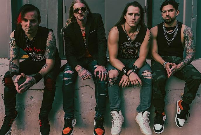 Four band members kneel at the stage edge as a packed crowd cheers behind them under the spotlights.