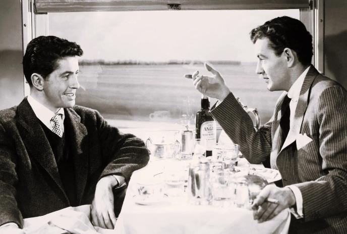 Two men in suits talk across a train dining table set with bottles, cups, and metal dishes