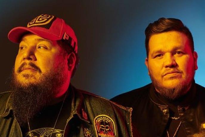 Two members of the band The Halluci Nation stand side by side against a gradient blue background.