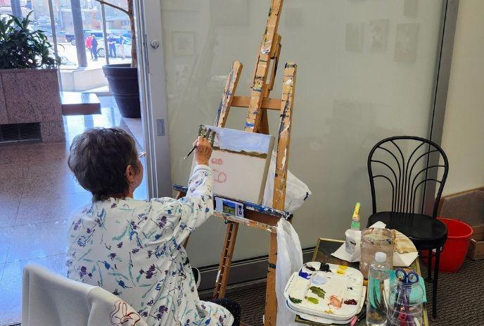 Artist painting while wearing patterned white outfit; canvas, palette, brushes, and bottles nearby.
