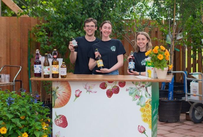 Three smiling staff members stand behind a decorated cider tasting bar at Heeman’s.