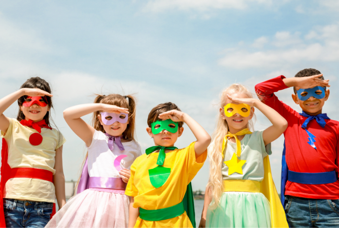 Five children in bright superhero costumes stand outdoors with hands raised as they look ahead.