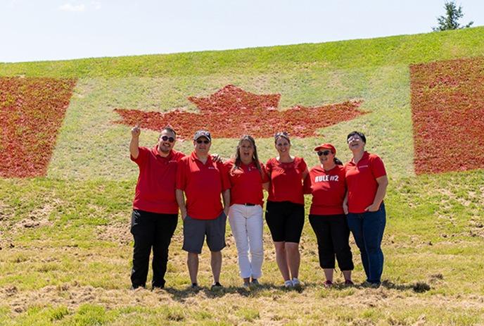 A group of six people in red shirts stand in front of a hill decorated with the Canadian flag using flowers or plants.