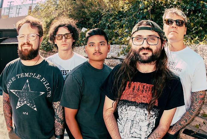 Hot Mulligan band poses with neutral, relaxed expressions in t-shirts against a white wall and a textured stone pillar.