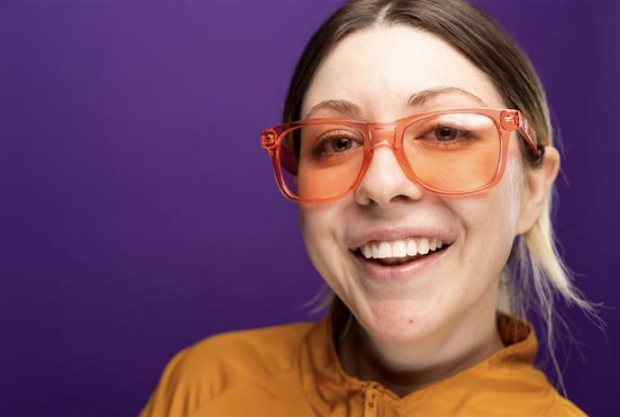 Jessie Johnson smiling for the camera in front of a purple background, wearing orange sunglasses.