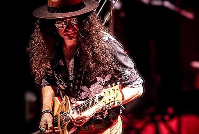 Musician with curly hair plays guitar onstage in starry shirt, hat, and shades under red stage lights.