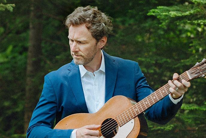 Ian Sherwood holding an accoustic guitar, with green pine trees in the background.