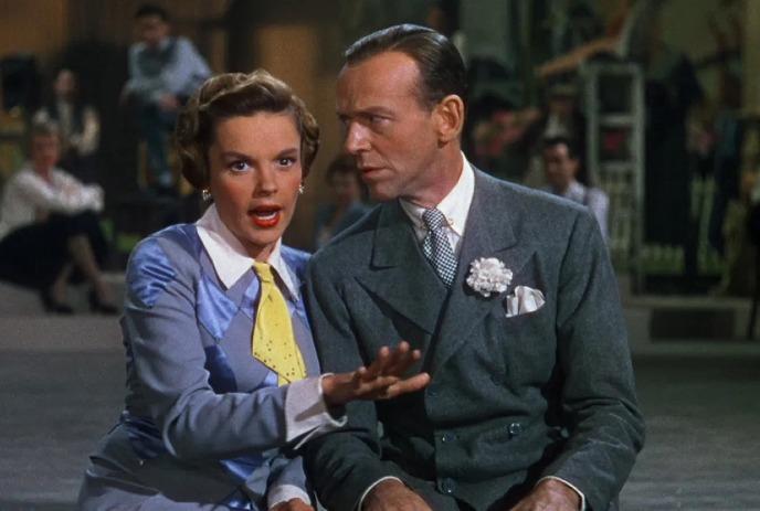 Two people sit close as one speaks with animated warmth while the other listens in a busy stage setting.