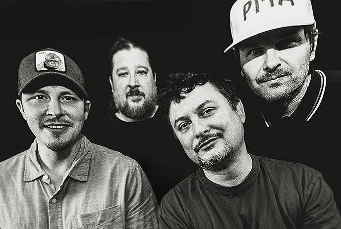 Four men of Illscarlett are standing together in a black and white image. Two of them are wearing caps.